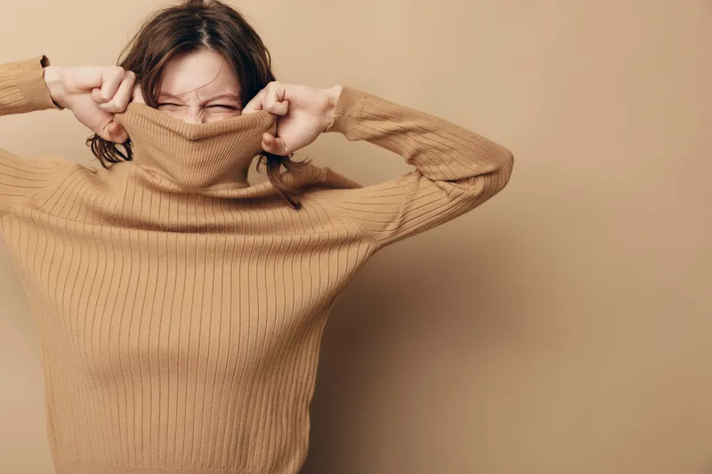 Young woman in beige sweater pulls collar over her face with closed eyes, playful expression on beige background. Concept of cozy clothing, autumn fashion, and casual style.