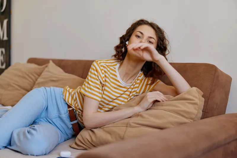 A young woman lounges on a beige sofa at home, wearing a striped yellow t shirt and light jeans, with a casual contemplative mood and relaxed posture.