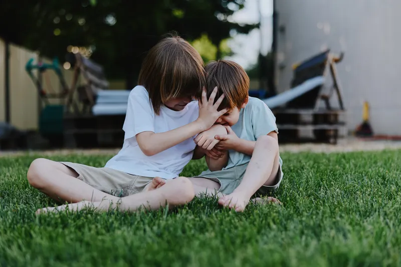 Two playful children laughing together on the grass, sharing a joyful moment in a sunny outdoor setting with a blurred background of greenery and wooden structures.
