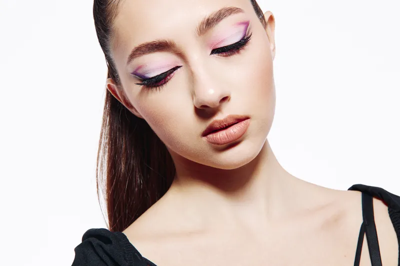 Close-up of a young woman with creative purple and pink artistic eye makeup looking down softly against a white background. Beauty and fashion concept with modern style.