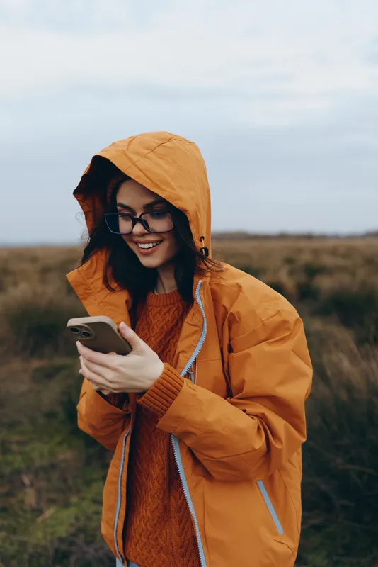 Woman lifestyle outdoors, smiling in orange hooded jacket, wearing glasses, holding smartphone, standing in autumn field, cloudy sky, candid portrait, warm casual fashion, natural light