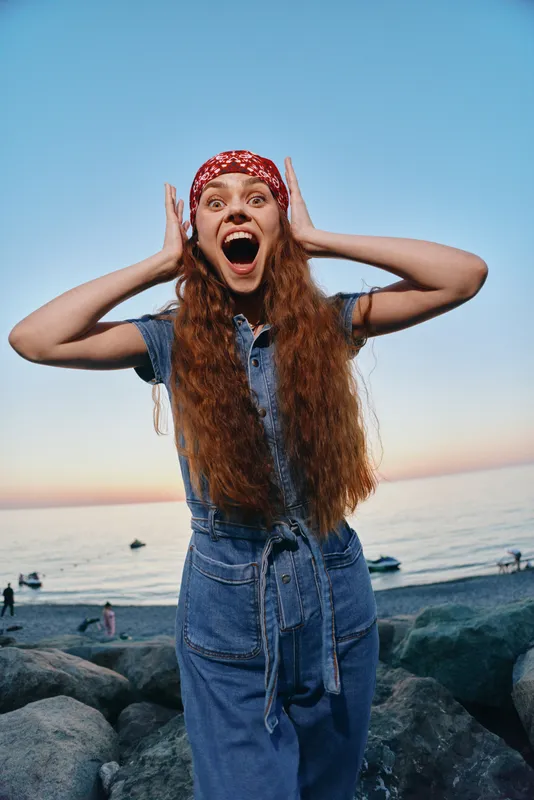 energetic woman in modern boho-western denim street style with long red hair and bandana, captured in vibrant film color near rocky beach at sunset