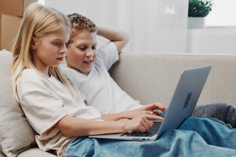 Two children sitting on a sofa using a laptop together in a bright living room. They are focused on the screen, sharing an educational or entertainment moment at home.