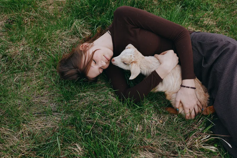 Goat, kid, woman resting outdoors on grass, gentle cuddle moment, peaceful countryside vibe, soft lighting, natural colors, simple lifestyle scene, emotional bond, serene mood