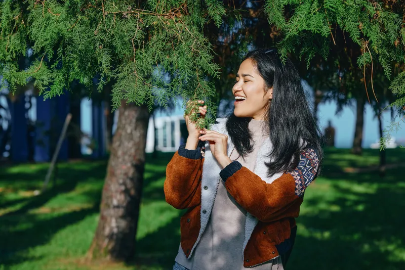 Woman, nature and outdoors scene with a smiling female in a brown and gray jacket gently holding a branch beneath dense green foliage in a sunlit park during a peaceful afternoon breeze