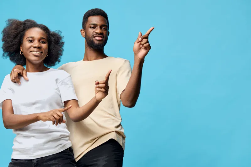 Happy young Afro American couple joyfully pointing at each other against a light blue background, dressed in casual white and beige shirts