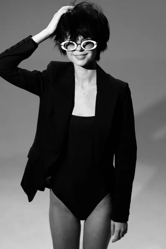 Black and white smiling woman in sunglasses and blazer posing with casual hairstyle in studio lighting.