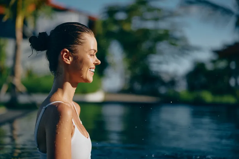 Smiling woman in a white swimsuit by calm water, side profile. Outdoor lifestyle scene highlighting relaxation, wellness, and summer leisure in warm sunlight.