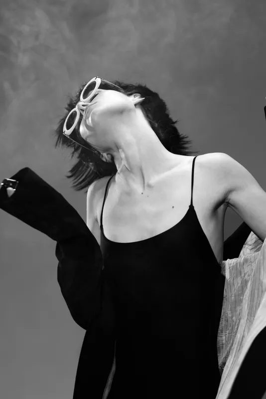 Black and white portrait of stylish woman in sunglasses posing with elegant expression and modern outfit in outdoor setting.