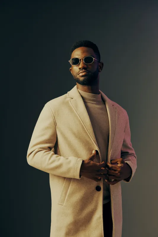 Stylish African man in beige coat and sunglasses, exuding confidence against a minimalist dark backdrop, perfect for fashion and lifestyle themes