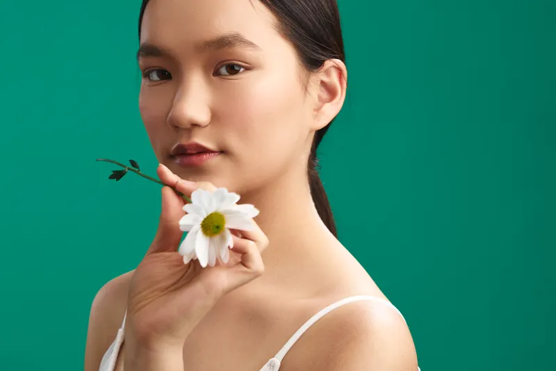 asian woman with a calm expression holds a white flower near her chin against a vivid green backdrop, creating a natural portrait that emphasizes softness, balance, and contemporary beauty.