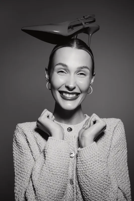 Smiling Caucasian woman with fair skin wears knitted jacket and balances high heel shoe on head. Fun joyful studio portrait concept.