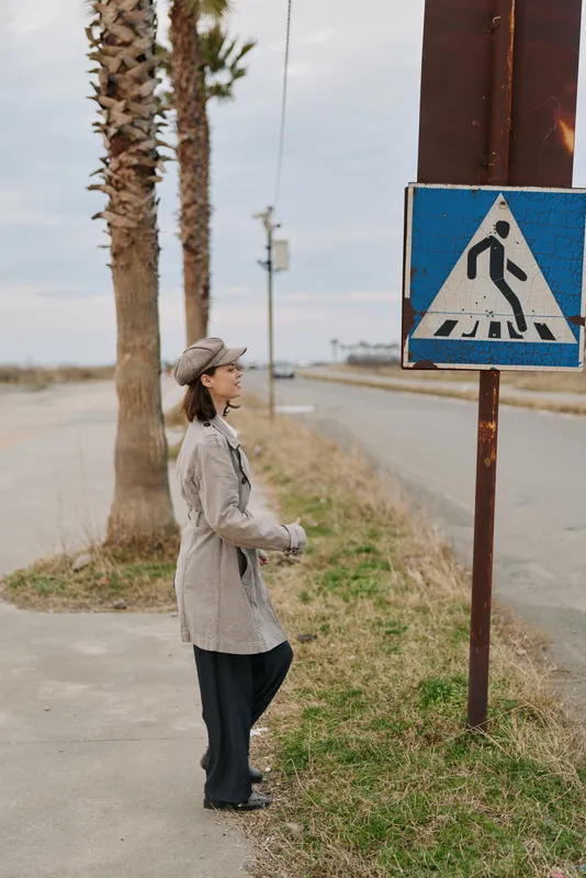 Woman in coat and hat stands near pedestrian crossing sign on roadside with palm trees and cloudy sky. Concept of urban lifestyle, travel, and casual fashion outdoors.