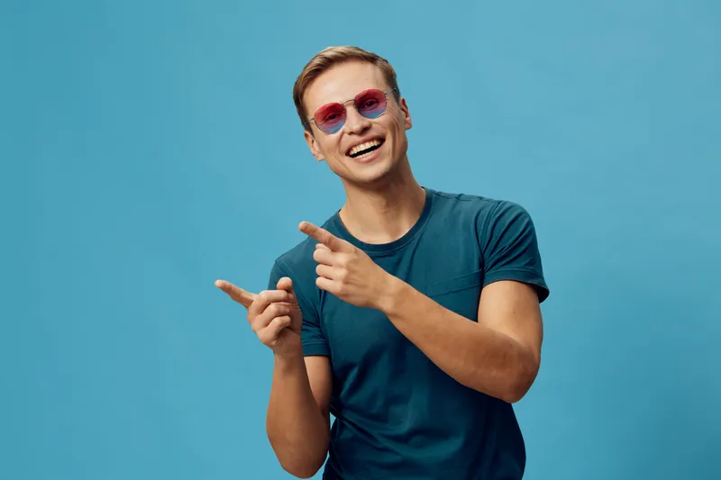 Smiling happy young caucasian man wear blue t-shirt red sunglasses pointing fingers isolated on blue background digital nomad lifestyle concept