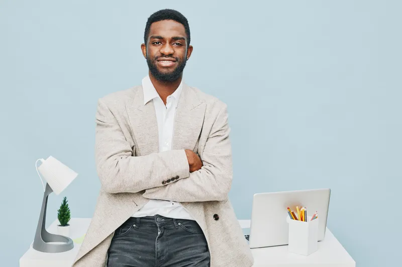Confident smiling man with dark skin, casual beige blazer and white shirt, standing with arms crossed at modern white desk, minimal office background, professional lifestyle concept