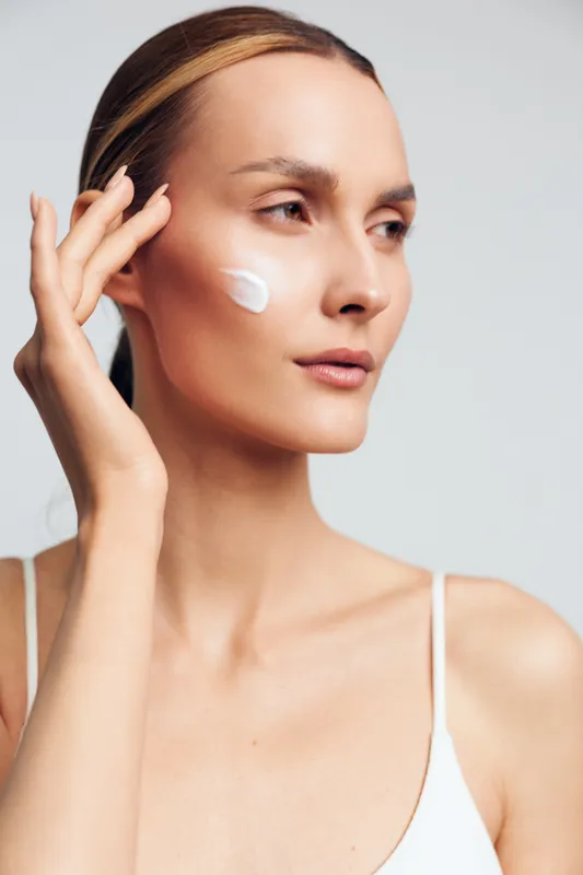 Woman applying face cream, close up on clean skin, soft studio lighting, she smooths a white moisturizer on her cheek, concept of skincare, wellness, self care, beauty routine, calm expression