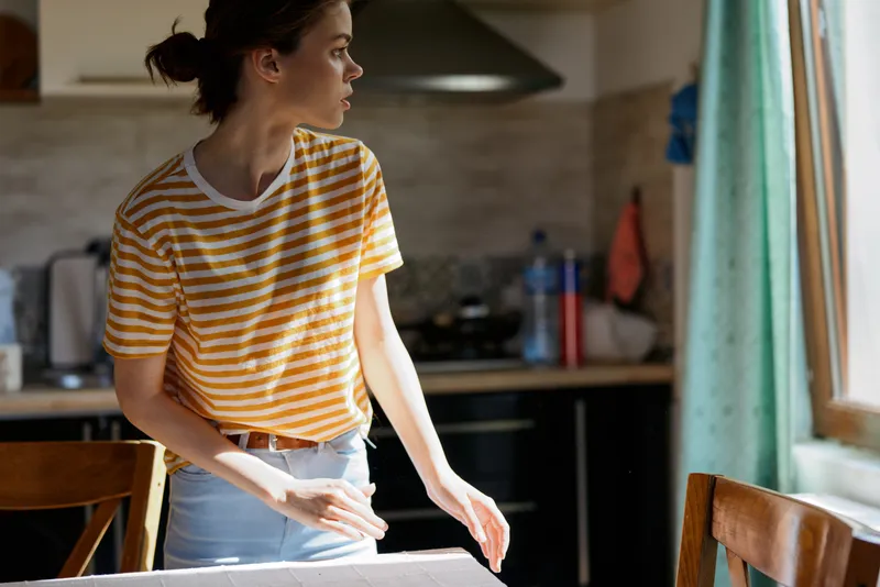 Woman in a striped shirt stands in a home kitchen, looking toward a sunlit window, capturing a casual morning scene and everyday domestic moment, relaxed and natural
