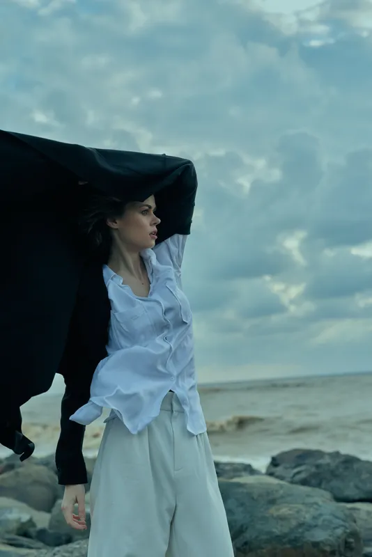 Wind sweeps across a rocky coast as a woman holds a jacket over her shoulder, standing by the sea beneath a moody sky, embodying outdoor style, independence and rugged atmosphere.