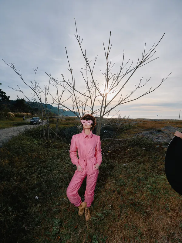 Fashion portrait outdoors with a pink jumpsuit worn by a model beside a leafless tree, open landscape and soft light creating a bold, modern style in a natural setting.