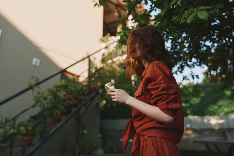 Woman in a red dress using a phone outdoors, capturing a casual lifestyle moment in bright daylight. Sunlit greenery creates a warm, relaxed mood and everyday charm, emphasizing contemporary living