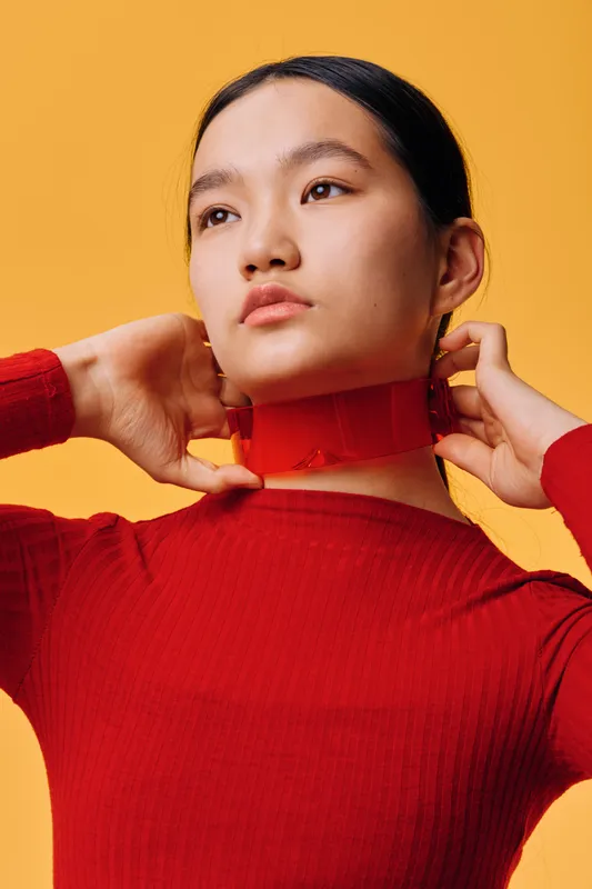 Young woman in red turtleneck sweater adjusting collar with hands, portrait on yellow background. Asian female model with smooth skin and natural makeup looking away thoughtfully.