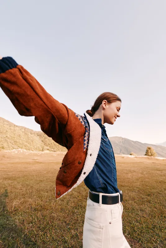 Woman in jacket in a field outdoors, portrait with smile and sense of freedom in the countryside. Happy nature lifestyle, denim shirt and warm coat captured in joyful motion.