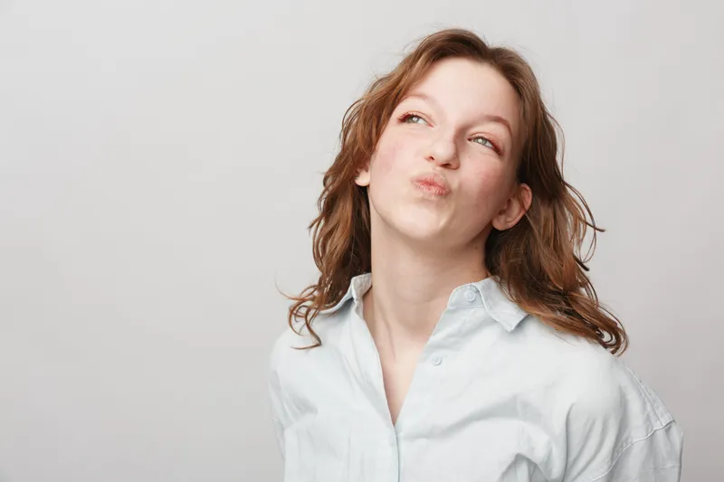 Young woman with wavy hair wearing white shirt makes playful face looking sideways on light background. Concept of fun, youth, casual style, and carefree attitude in studio portrait.