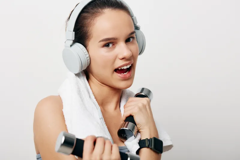 Woman in a gym setting, fitness enthusiast, lifting dumbbells with a towel draped over her shoulders, wearing large headphones and a determined expression during a focused workout.