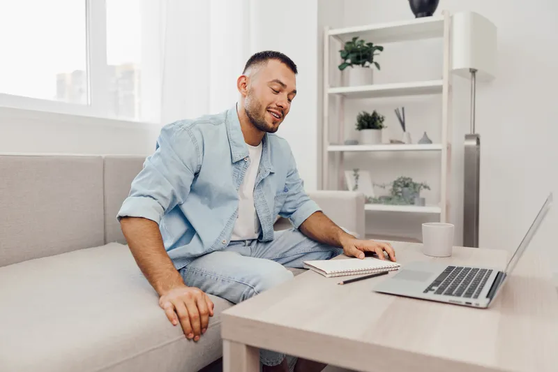 Smiling man engaged in remote work, sitting comfortably in a modern living room, surrounded by minimalistic decor and soft natural lighting, showcasing productivity and relaxation.
