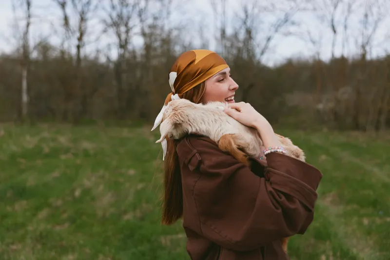 Smiling woman in a brown coat hugs a goat kid in an open field, warm scarf, autumn light, peaceful mood, lifestyle moment, animal companionship, gentle care, natural warmth radiates from the scene