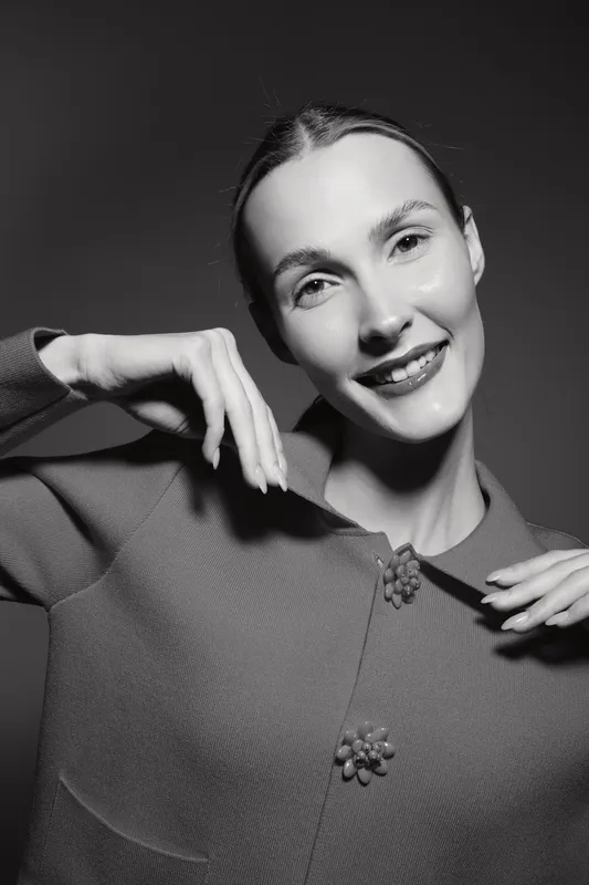 Young white woman with fair skin smiles posing in textured jacket against dark background. Studio portrait with soft lighting emphasizes natural beauty and elegance.