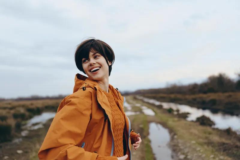 Woman lifestyle outdoors, smiling as she hikes along a river path, wearing an orange jacket and scarf. Scenic landscape with cloudy sky, soft focus, calm travel mood on a breezy day.
