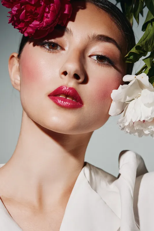 Close-up of a young woman with glossy red lips and dramatic makeup featuring cheek contour and floral accessories. Beauty and fashion creative concept with vibrant style.