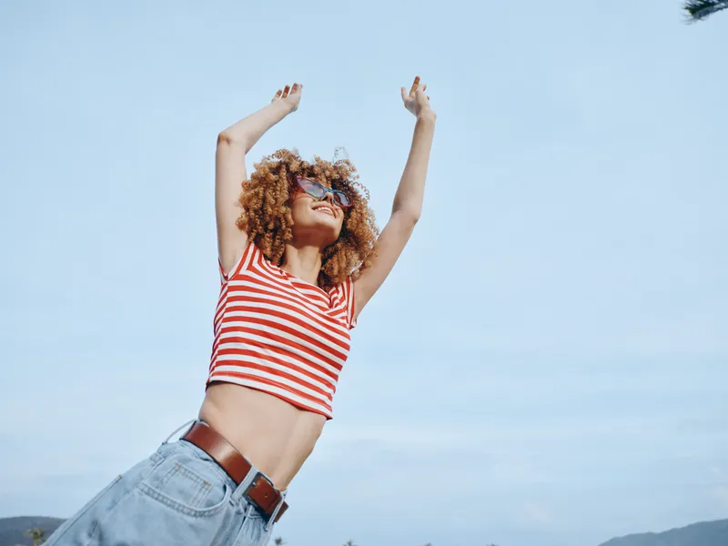 Young woman with curly hair wearing a striped crop top and denim jeans raises her arms against a clear blue sky, radiating freedom, joy, and sunny energy.
