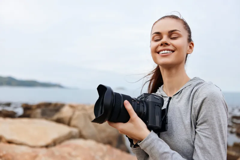 Happy young woman holding a camera by the seaside, showcasing her love for photography and natural beauty with a vibrant coastal backdrop