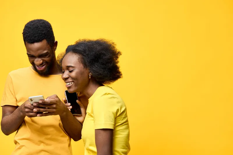 Happy young African couple enjoying their time with smartphones against a vibrant yellow background, showcasing joy and connection in a playful atmosphere