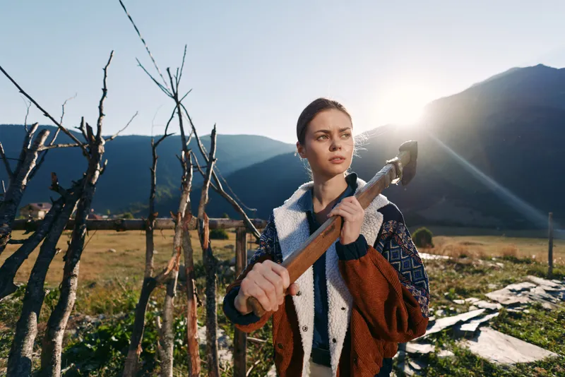 Woman with axe near a wooden fence in rugged mountains on a small farm at sunrise, rural portrait capturing determination and rustic outdoor lifestyle with sunlight on meadow.