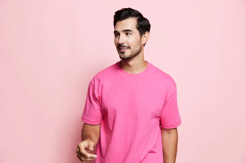 Stylish smiling man wearing a bright pink t-shirt looks off to the side while gesturing with his hand against a solid pastel pink background. Modern lifestyle concept.