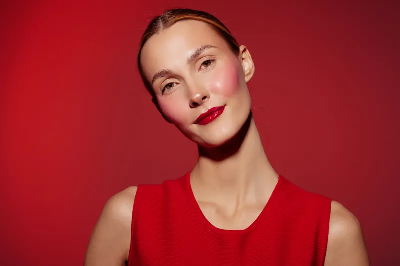 Portrait of a young woman with red lipstick and red dress on red background. Studio lighting highlights her clear skin and confident expression. Concept of beauty and fashion.