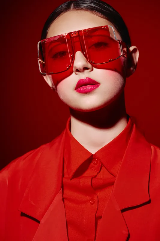 Stylish young woman with vibrant red sunglasses and red lipstick stands against red background creating bold beauty and fashion creative concept in studio lighting.