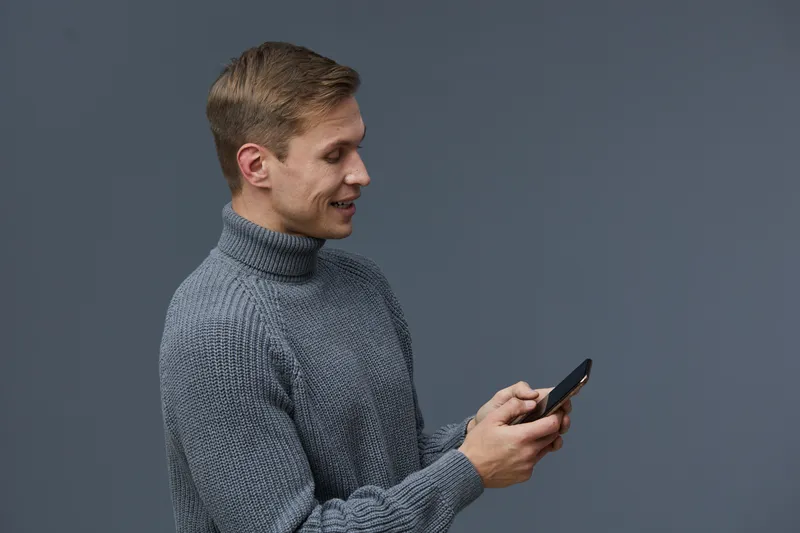 Smiling young caucasian man wearing gray sweater holding smartphone texting chat with AI digital communication concept isolated on gray background studio portrait