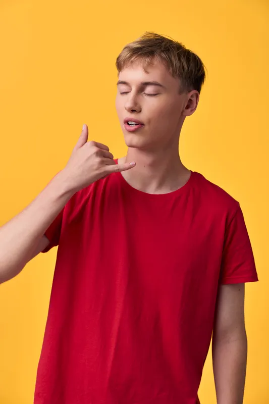 Teenage boy in a red shirt posing with a hang loose gesture, eyes closed, on a bright yellow backdrop, casual street style, youthful energy, playful mood, lifestyle photograph