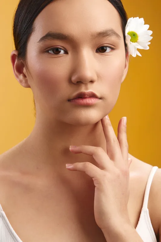 Asian woman is framed in a close up portrait with a white flower tucked behind her ear. She offers a calm, elegant gaze, conveying serene beauty and poise, with gentle lighting and soft skin tones.