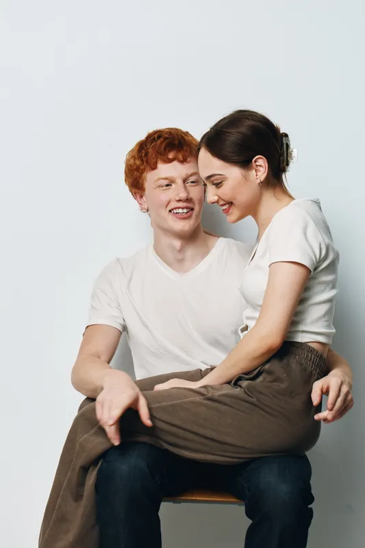 Young couple sitting together with affection on chair, smiling and enjoying moment. Man with red hair holds woman on lap in casual clothes against white background.