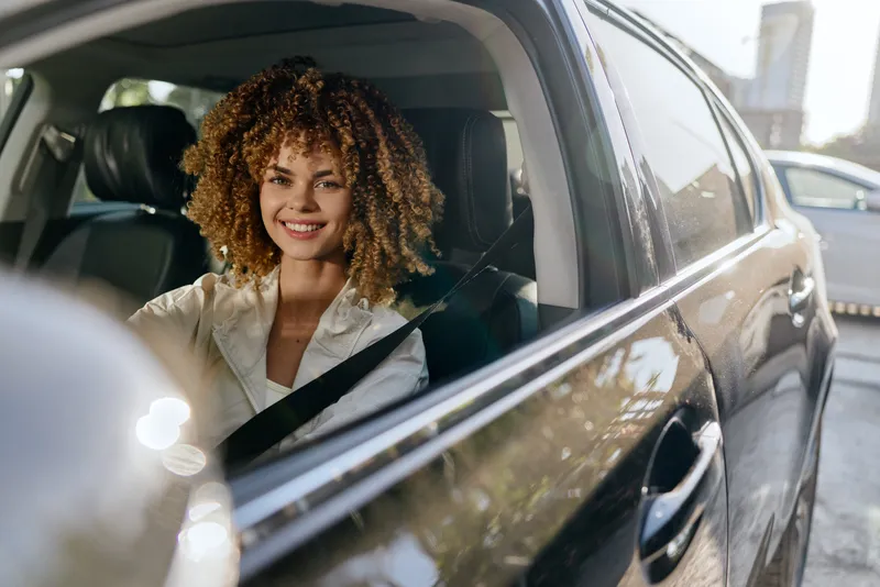 Smiling woman driving a car with curly hair wearing a white blouse, enjoying a sunny day in an urban environment with modern buildings in the background.