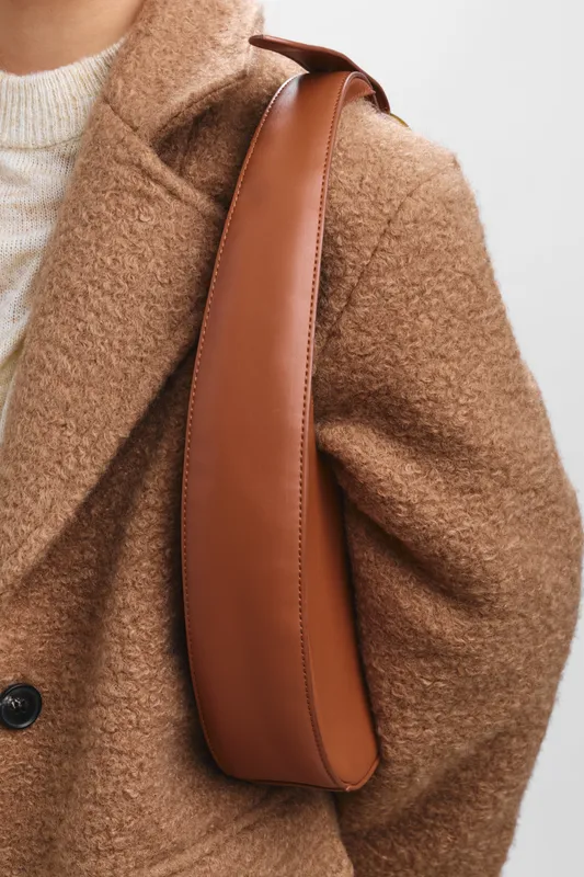 asian woman wearing a warm brown coat with a leather shoulder strap, standing in a minimalist setting, conveying chic urban style and quiet confidence with a sleek, modern silhouette.