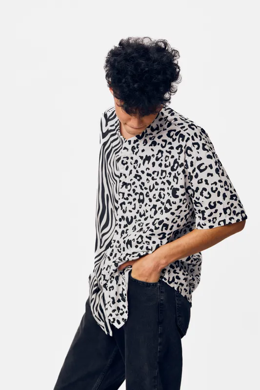 Young man wearing a trendy black and white animal print t-shirt with curly hair, looking down thoughtfully against a plain white background in casual style.