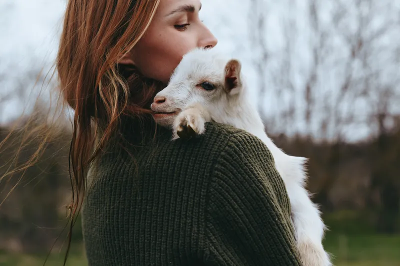 Lifestyle portrait of a woman outdoors, holding a goat kid in a cozy sweater, affectionate moment, intimate embrace, nature backdrop, soft light, calm breeze, warm colors, genuine connection, serene