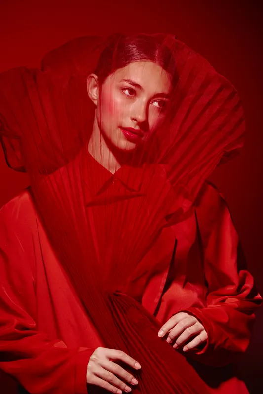 Young woman wearing avantgarde red dress and exaggerated collar made of pleated fabric stands against red background. Artistic fashion and creative beauty concept.