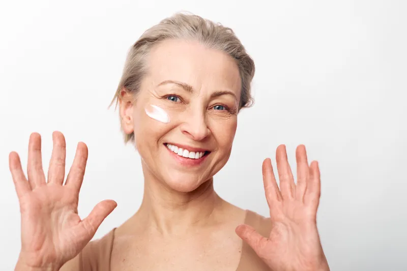 Smiling mature woman with short hair applies white face cream on cheek, showing hands in front of white background. Concept of skincare, beauty, antiaging, and self care routine.